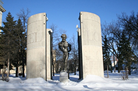 Figure fantaisiste en bronze de Louis Riel centrée au milieu de quatre hautes colonnes de ciment en quart de cercle, sur lesquelles on peut lire LOUIS RIEL.